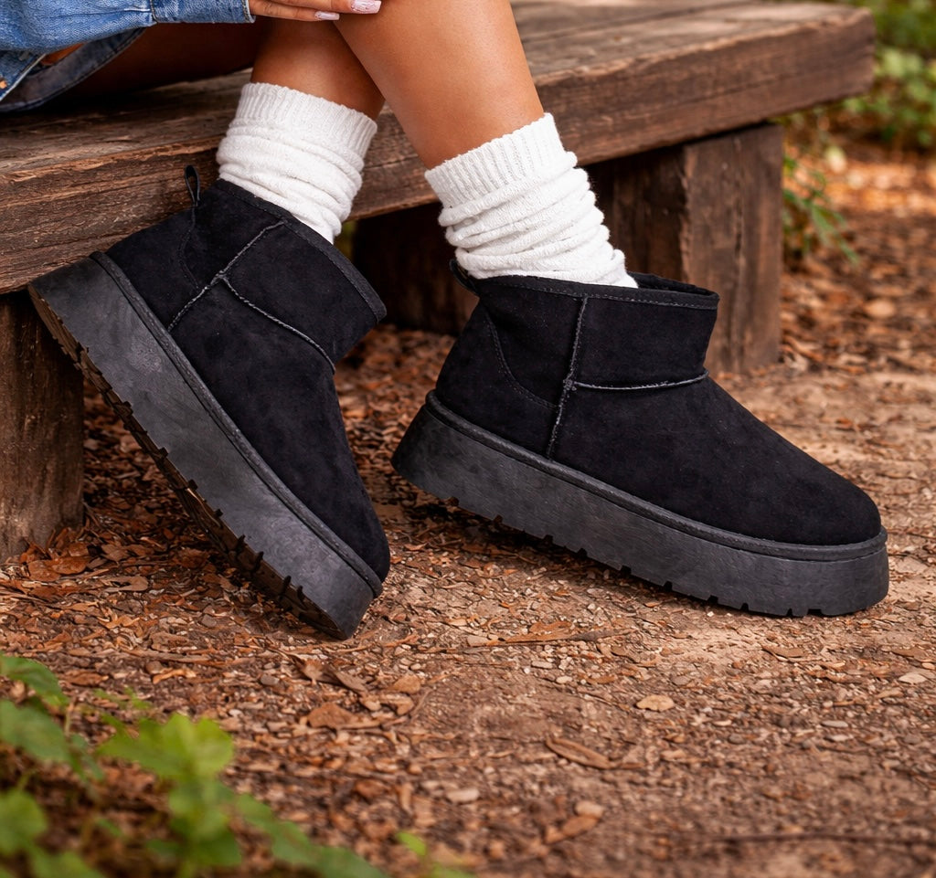 Black suede boots worn with white socks on a wooden bench.