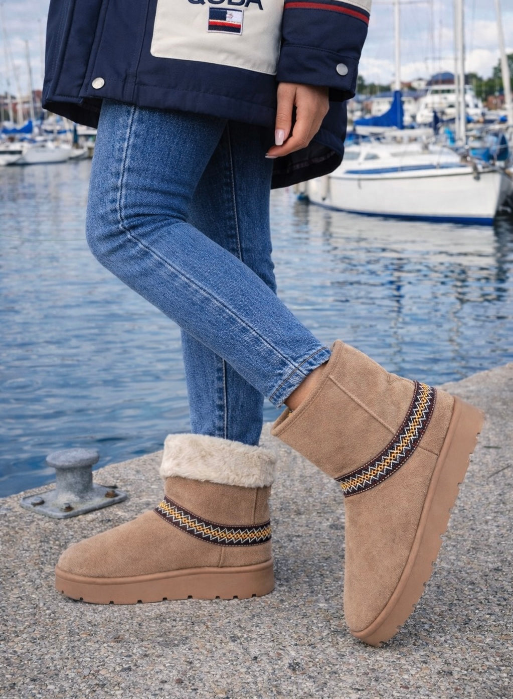 Person wearing tan suede boots with decorative straps by a waterfront with boats.