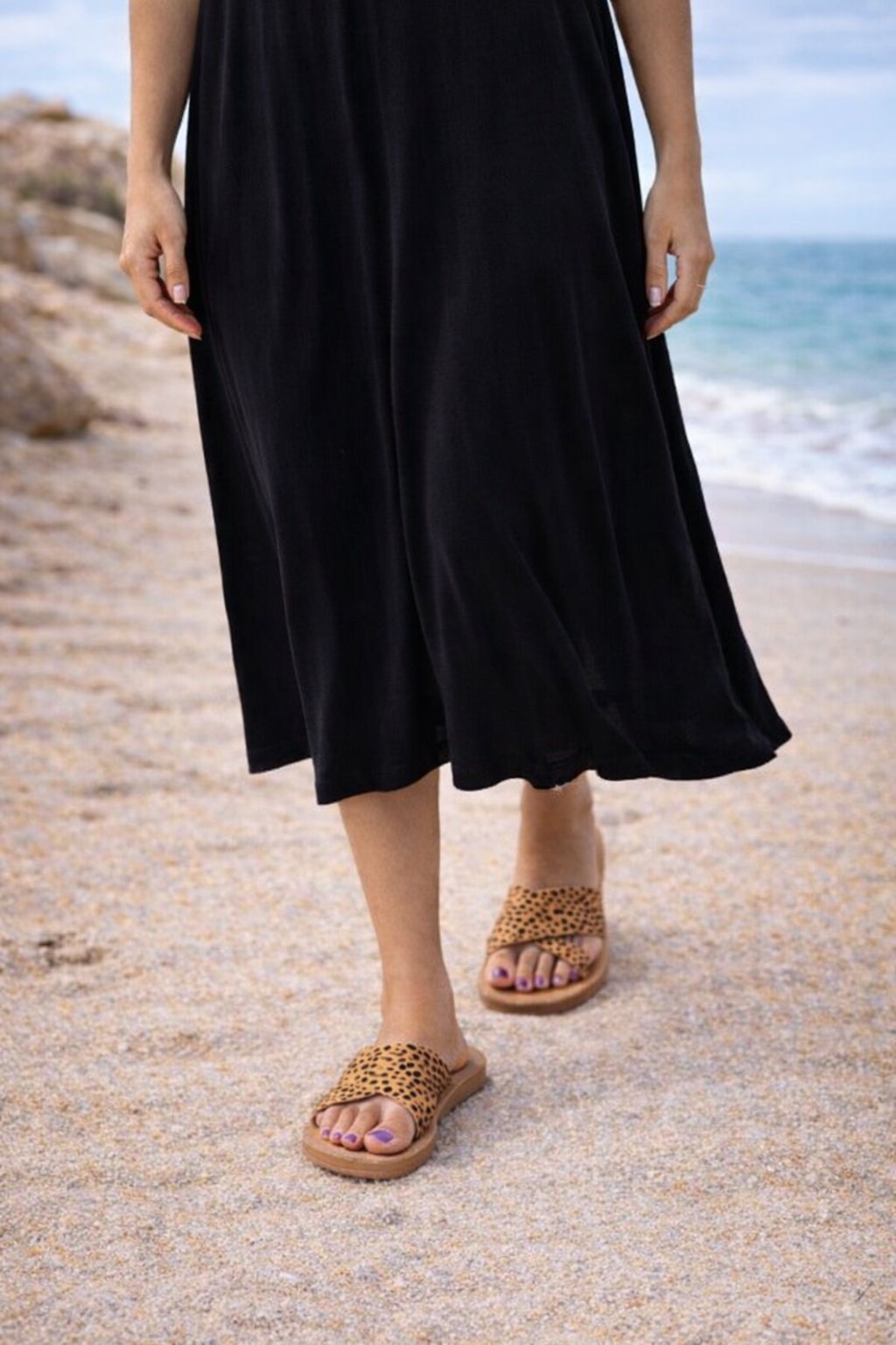 Person wearing a black dress and leopard print sandals on a beach.